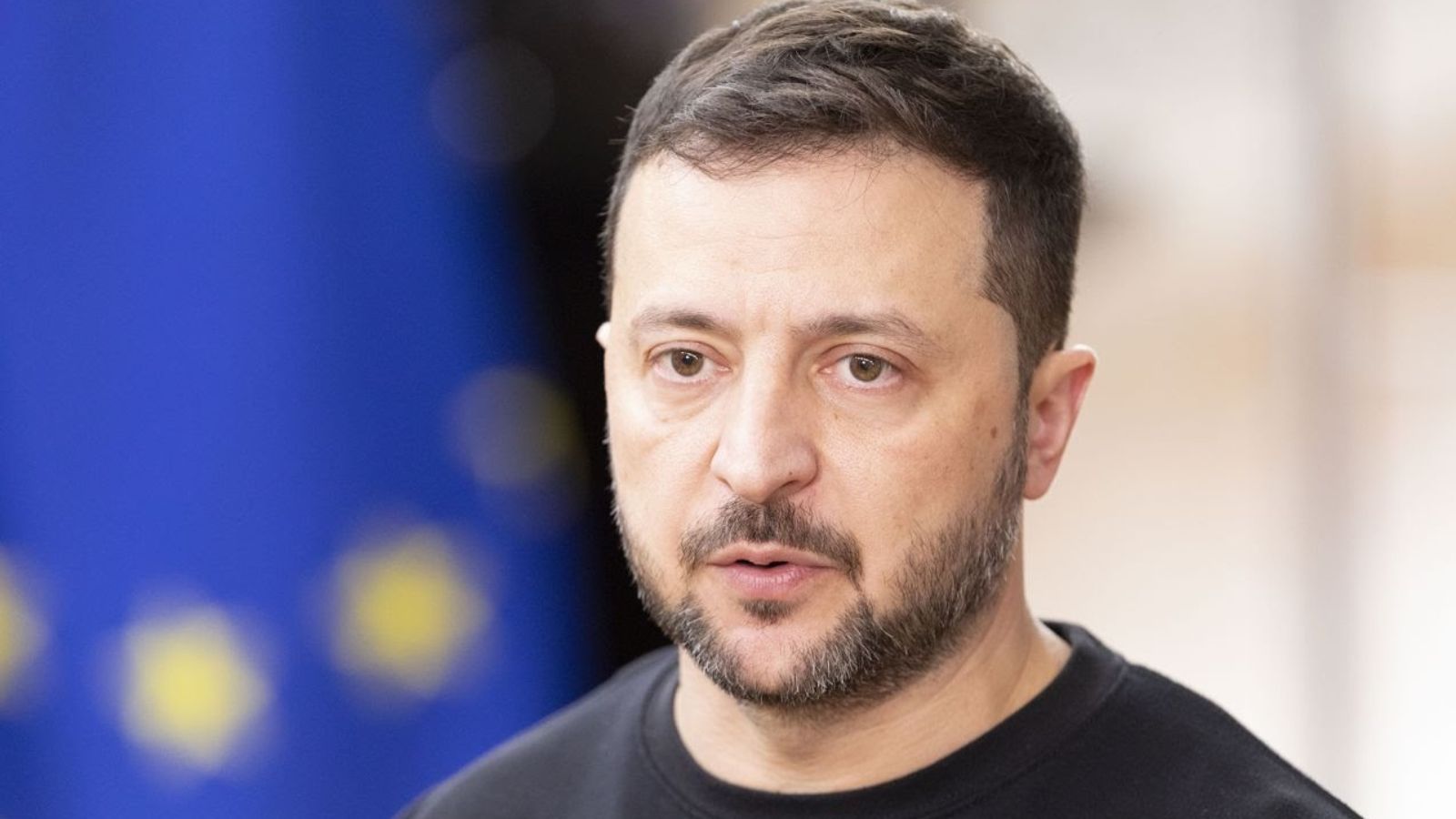 Портрет великим планом Президента України Володимира Зеленського із зосередженим виразом обличчя. На задньому плані розмитий прапор Європейського Союзу.
