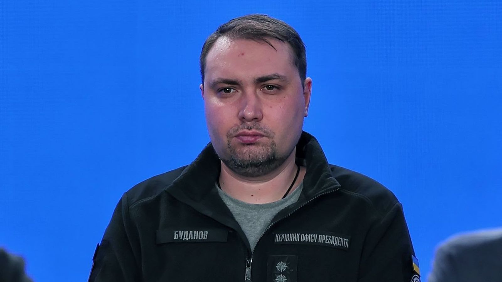 Portrait of Kyrylo Budanov against a bright blue background. He is wearing a black tactical fleece with a "Budanov" name patch.