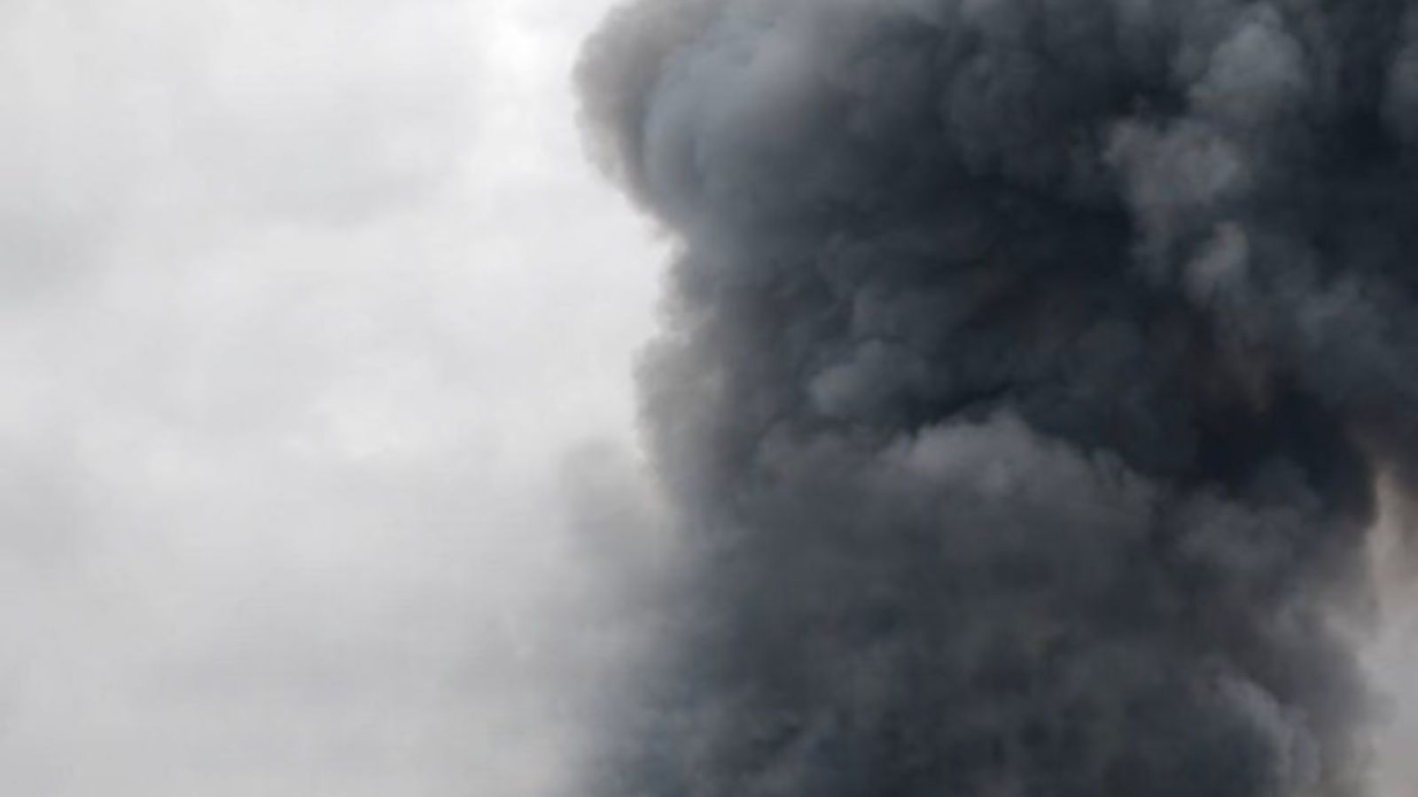 A pillar of thick dark gray smoke rises high into a cloudy sky, filling most of the frame.