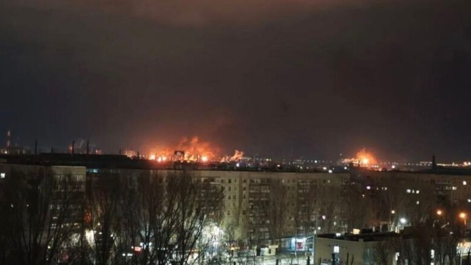 Panoramic night view of an industrial area of a city, with the glow and fire of a large-scale blaze visible on the horizon after an attack. In the foreground are the roofs of residential buildings.