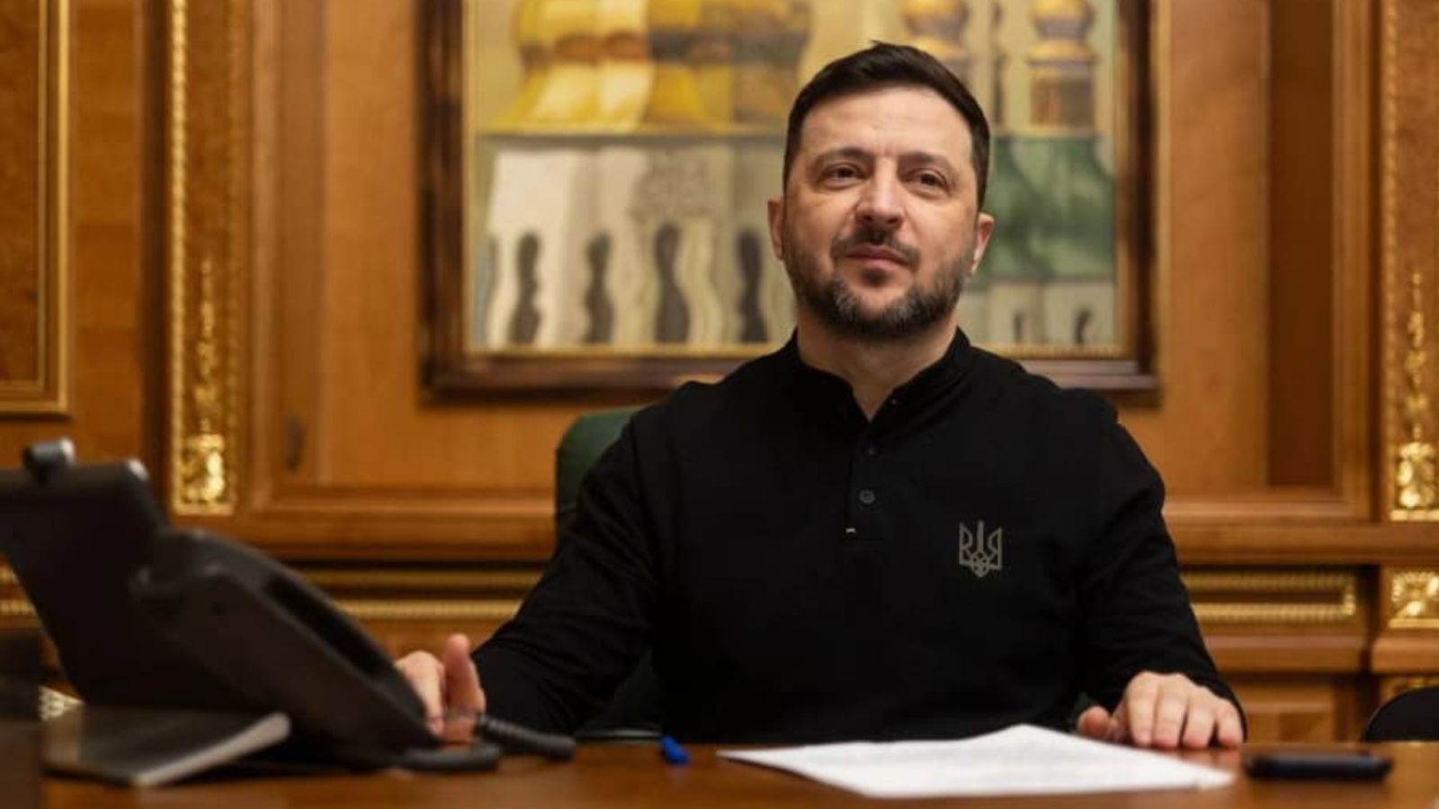 Volodymyr Zelenskyy in a black vyshyvanka is sitting at a large wooden desk in his office. He is looking directly at the camera, his hands resting on the table near some papers. Behind him are wooden wall panels, gold trim, and a painting of church domes. A desk phone is visible in the foreground.