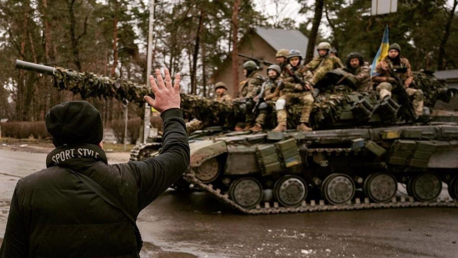 Цивільна людина зі спини махає рукою групі українських військових, які їдуть на броні танка по деокупованій території.