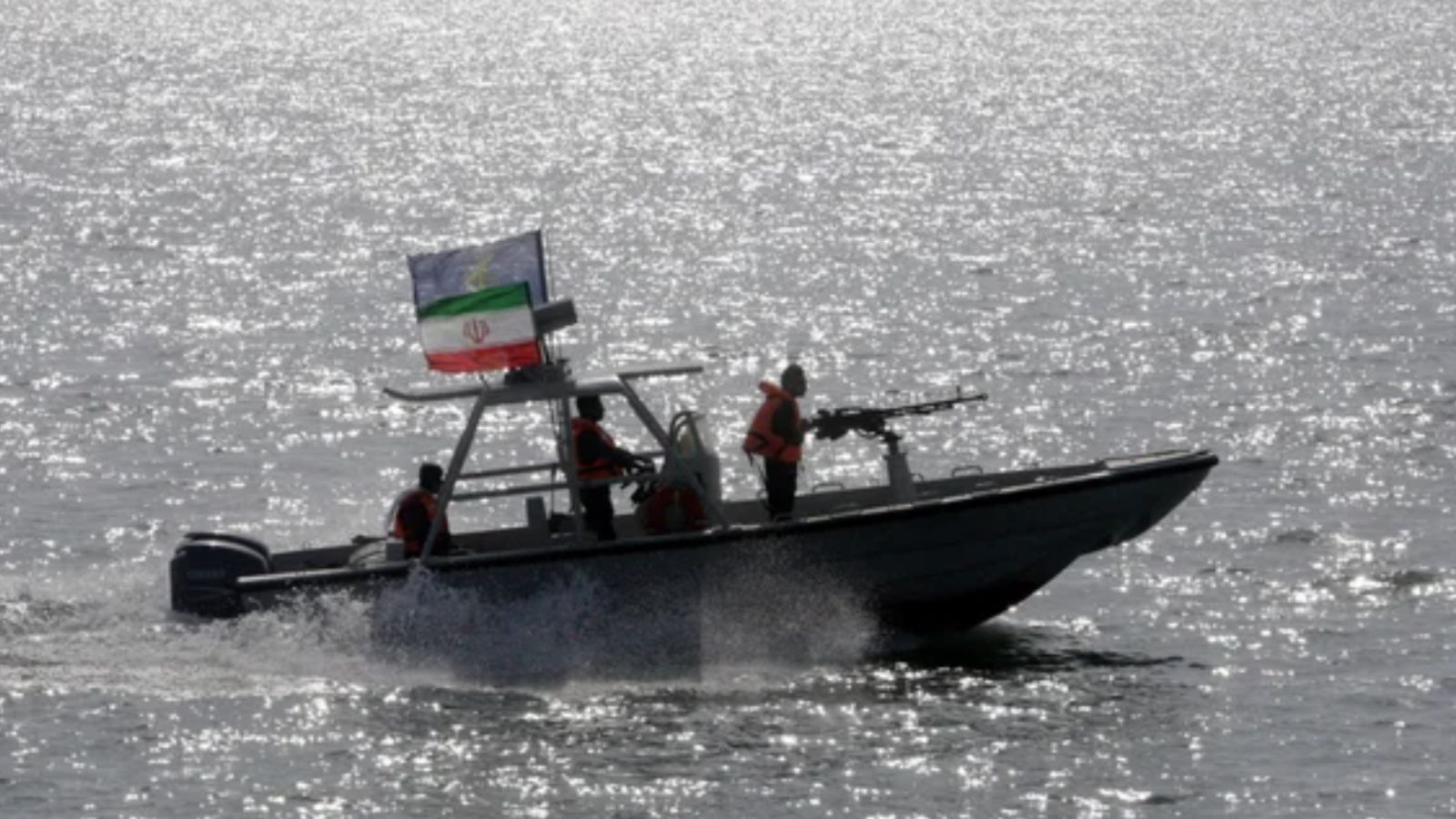 A photograph of a small, fast Iranian military patrol boat with two crew members aboard, moving across the water under bright sunlight. An Iranian flag is visible at the stern of the boat, and one of the military personnel stands by a machine gun.