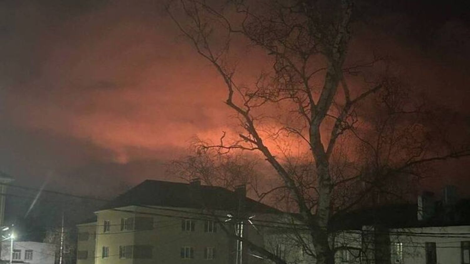 A view from a residential area showing the night sky lit up by a large fire following a drone attack in Kstovo. Silhouettes of buildings and trees are visible in the foreground.