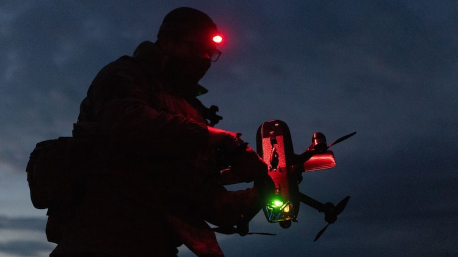 A soldier in the dark with a red headlamp on, holding a small drone with glowing status lights.