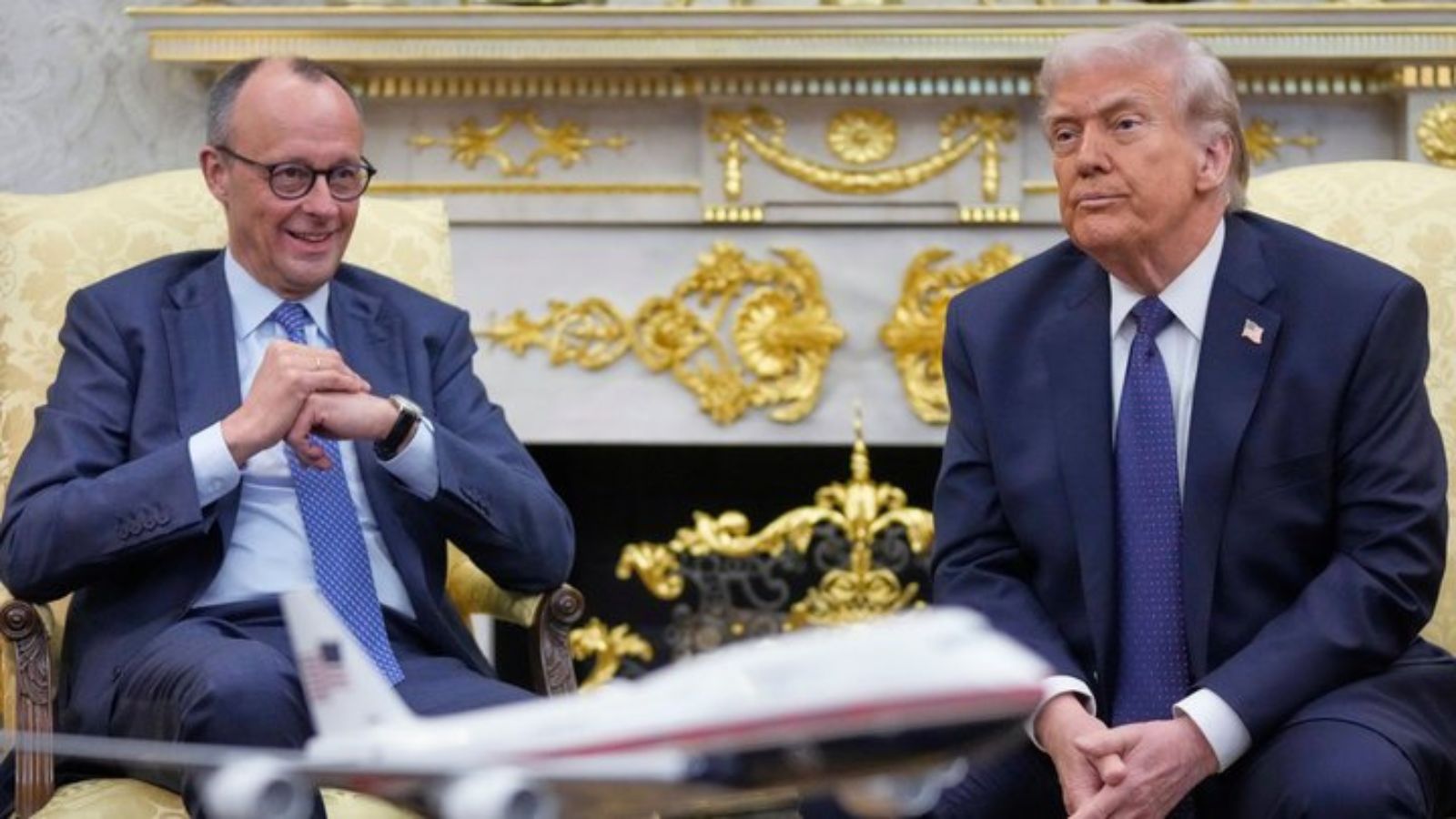 Donald Trump and Friedrich Merz are sitting in armchairs during an official meeting inside the White House. Merz is smiling with his hands clasped; Trump is looking away with a serious expression.