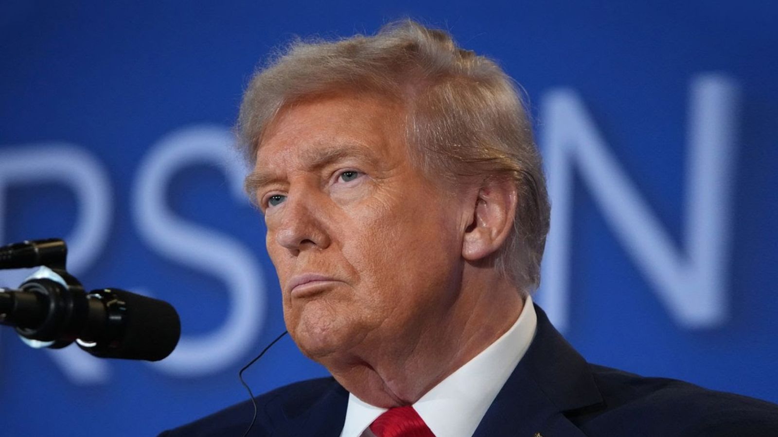 Дональд Трамп у навушниках дивиться убік, на задньому плані - розмитий напис.