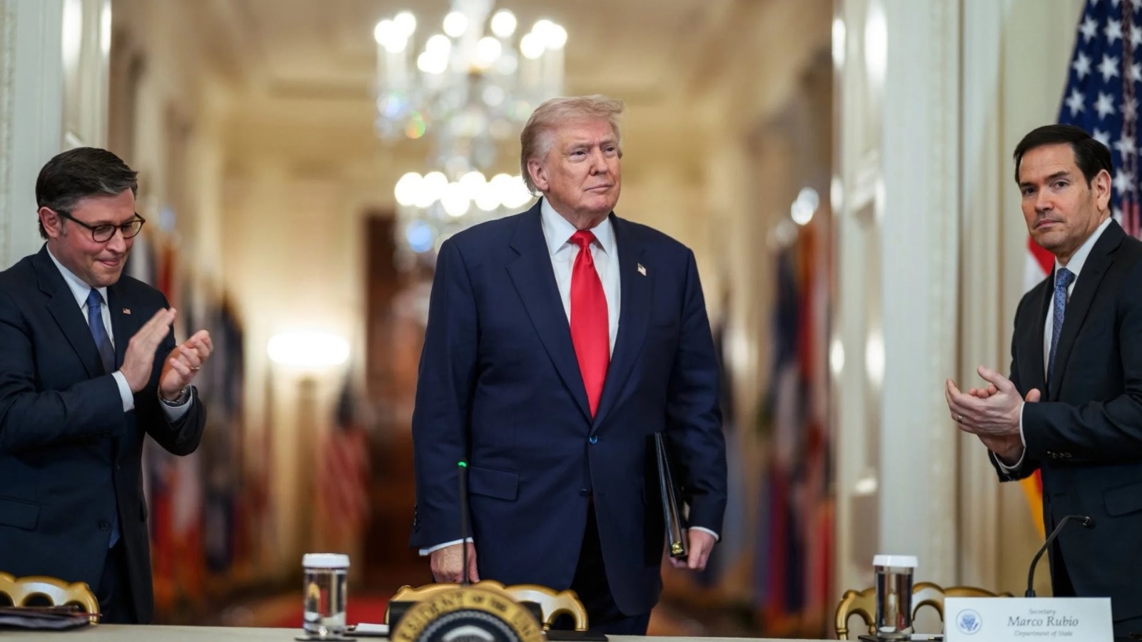 Donald Trump in the center of a White House hallway, clapping along with Mike Johnson and Marco Rubio standing beside him, with a nameplate for "Secretary Marco Rubio" visible on the table in front.