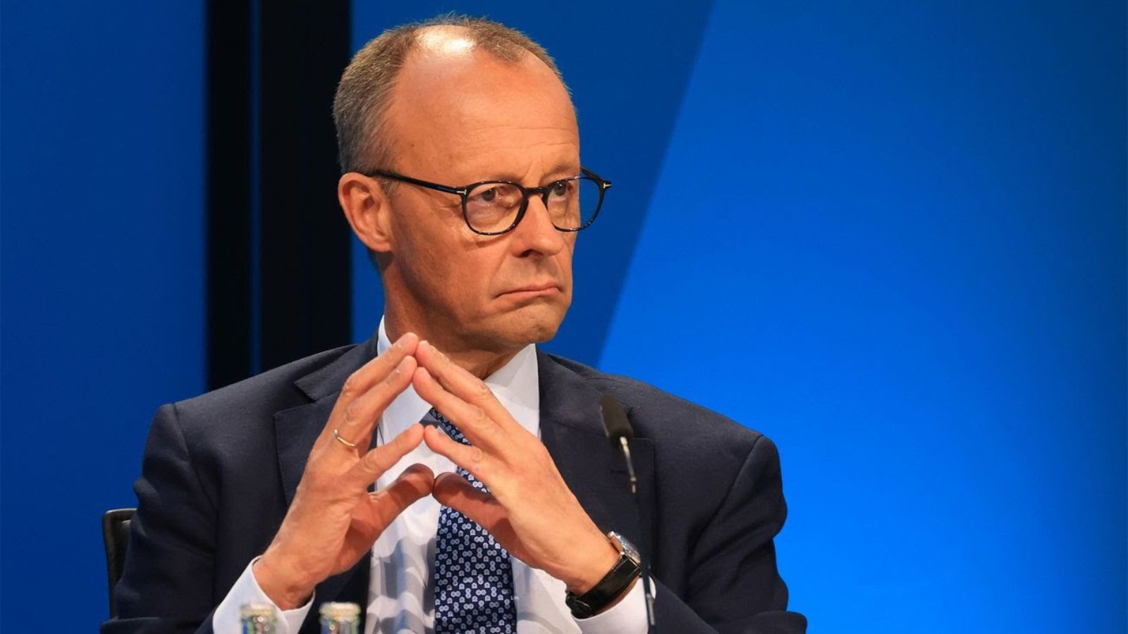 Friedrich Merz sitting at a desk with his hands clasped in front of him against a blue wall during an official event.