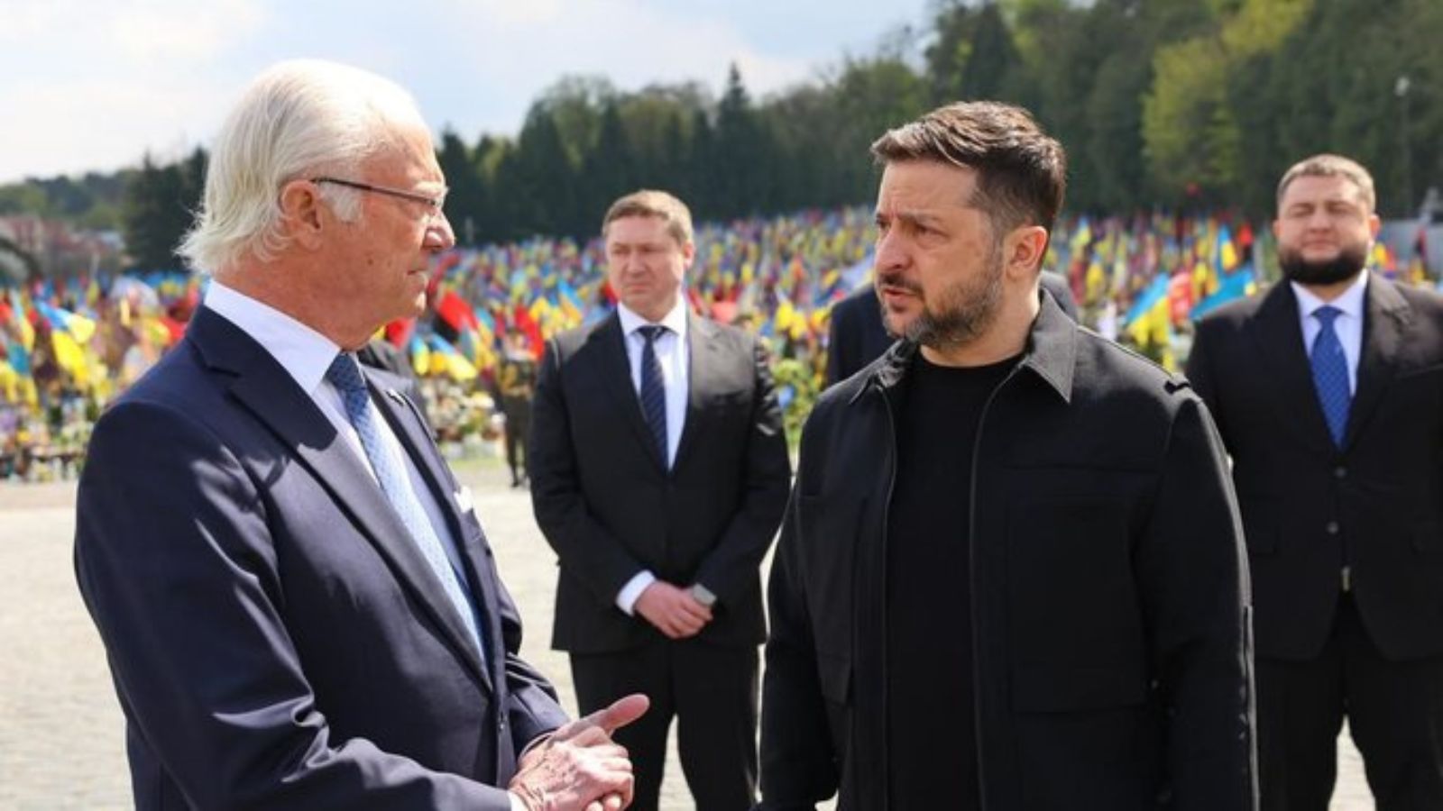 Volodymyr Zelenskyy and King Carl XVI Gustaf talking at the Lychakiv Cemetery in Lviv, with military graves and numerous Ukrainian flags visible in the background.