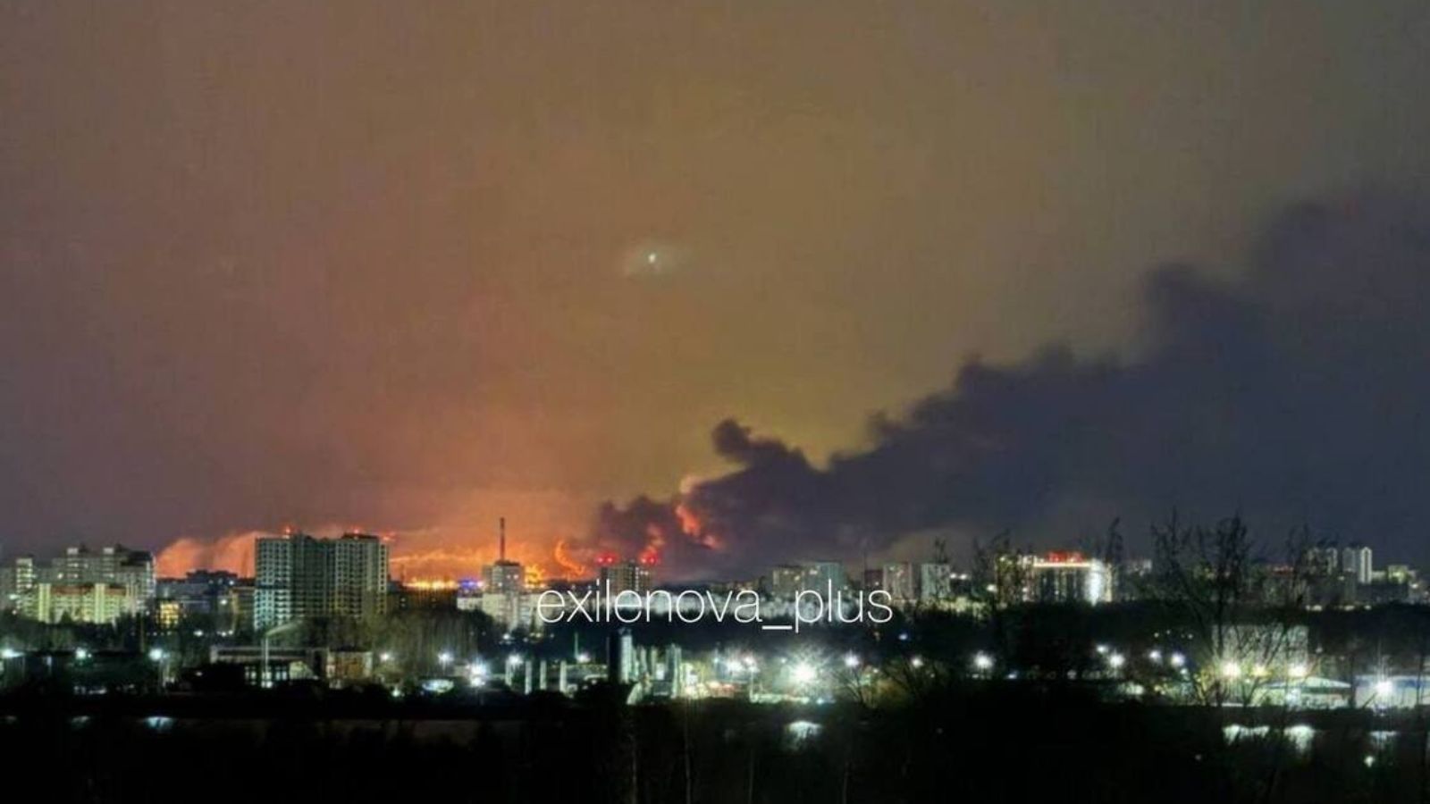 A nighttime city panorama showing a large fire and thick black smoke on the horizon behind residential buildings.