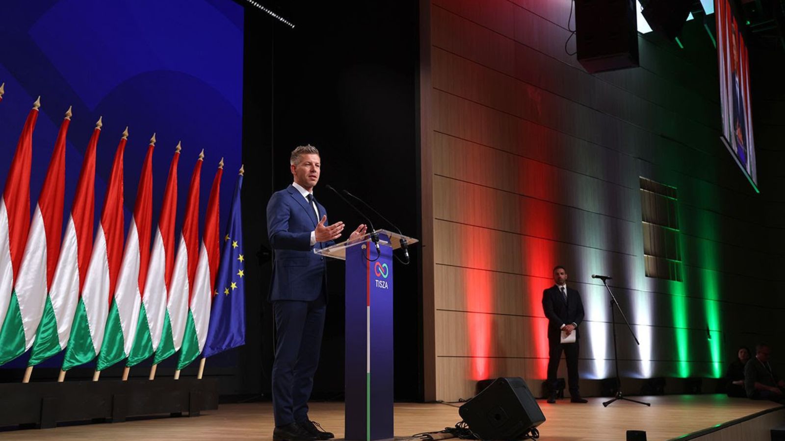 Péter Magyar, leader of the Tisza party, speaks behind a lectern on stage. To the left is a line of Hungarian and European flags. The back wall is illuminated in red, white, and green colors. The lectern features the Tisza logo.