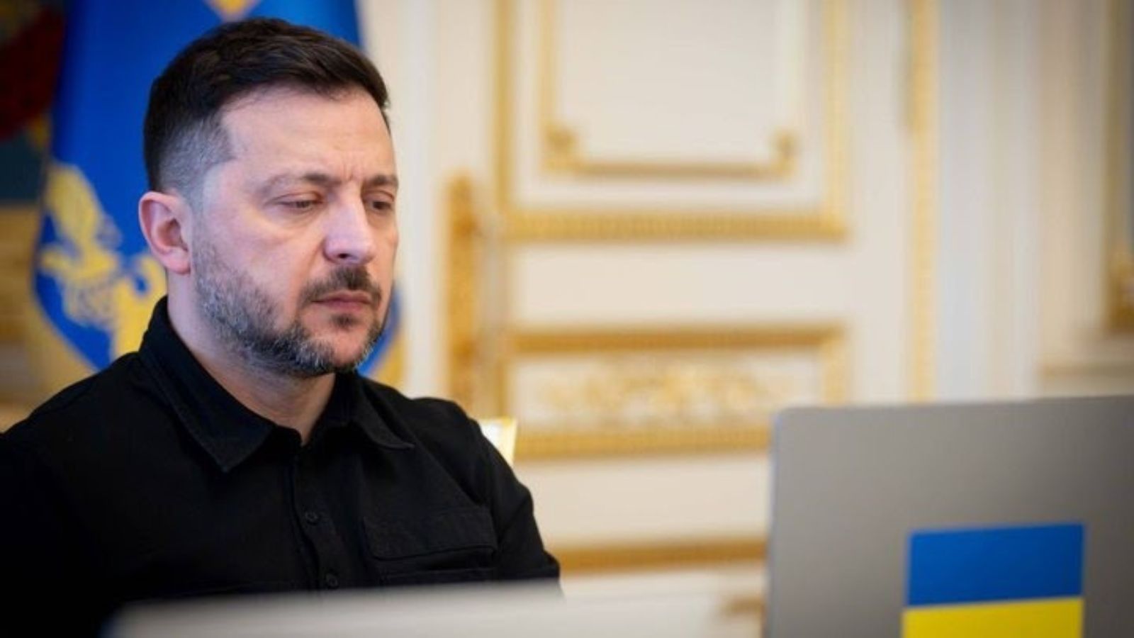 President of Ukraine Volodymyr Zelenskyy in a black shirt focuses on a laptop screen featuring a small Ukrainian flag sticker on the lid.
