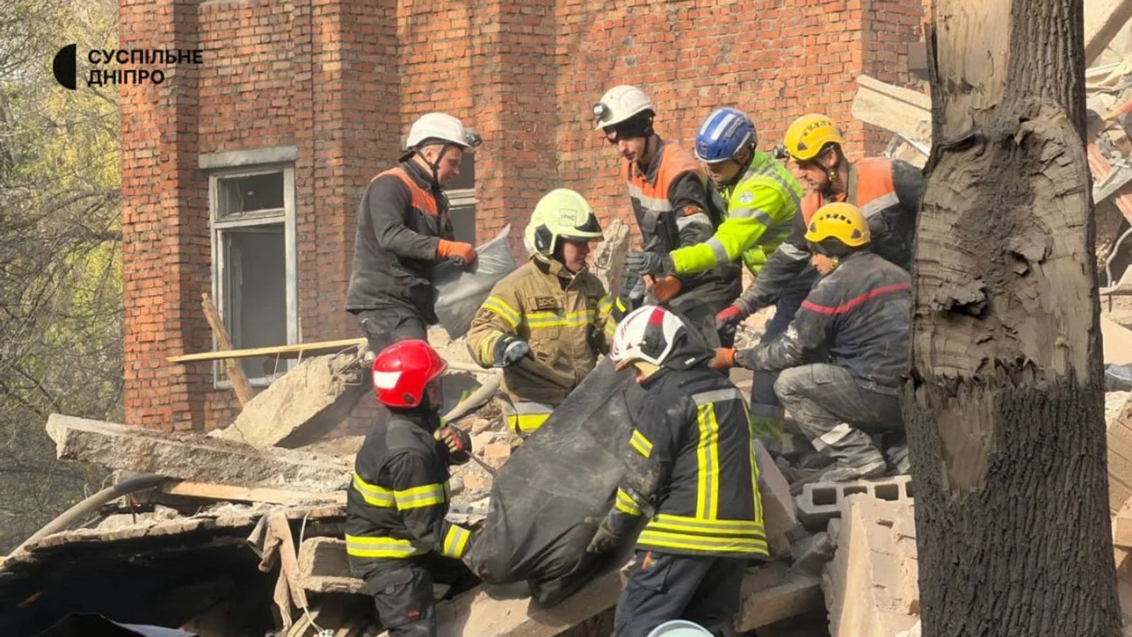 A close-up view of a group of rescuers wearing helmets of various colors (white, bright yellow, blue) working on the rubble of a destroyed building in Dnipro.