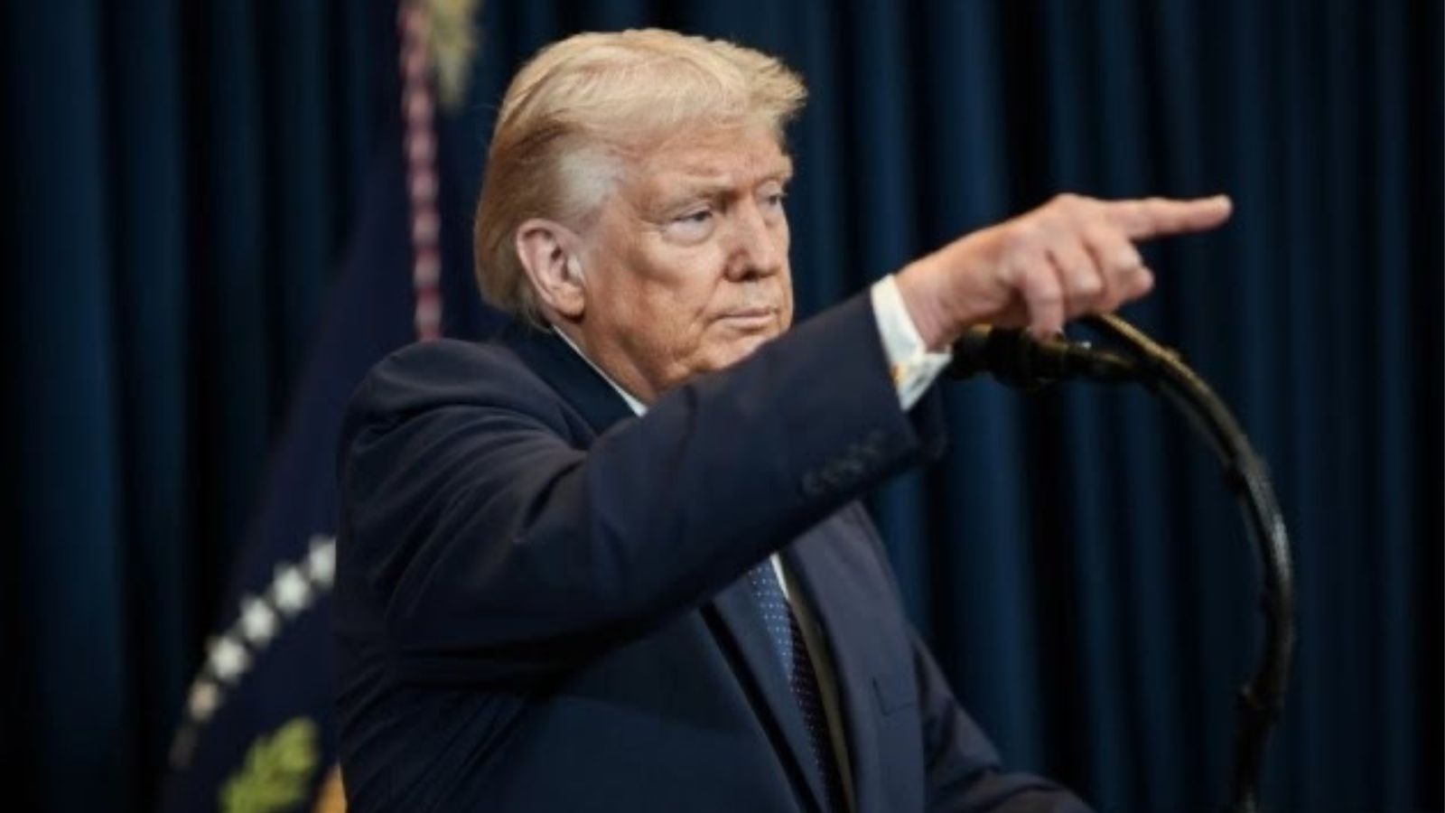 Дональд Трамп стоїть біля мікрофона на темному фоні та вказує пальцем вперед під час виступу.