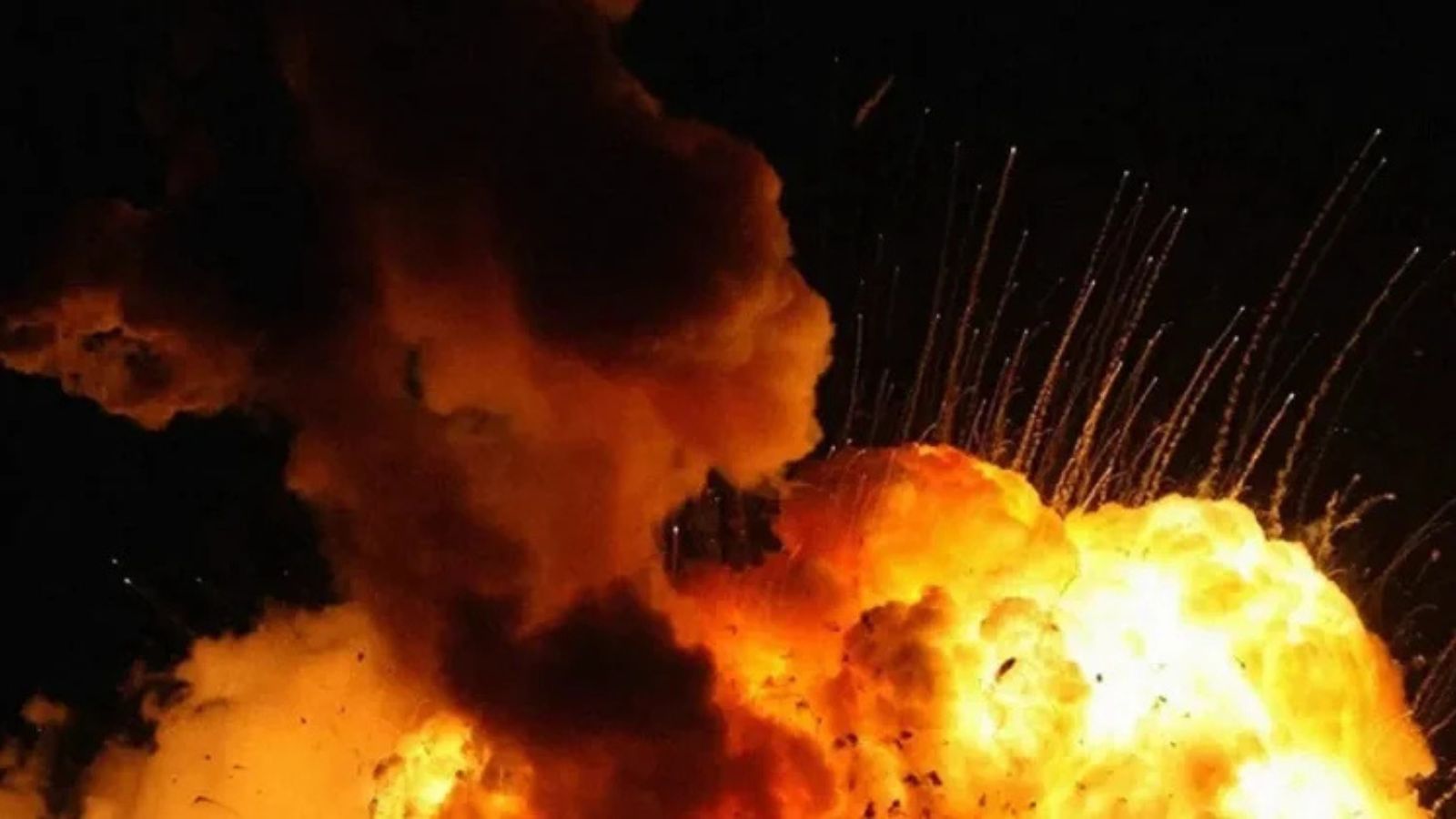 A massive nighttime explosion. A large smoke plume and bright orange and yellow fire with fiery sparks streaking upwards against the dark sky.