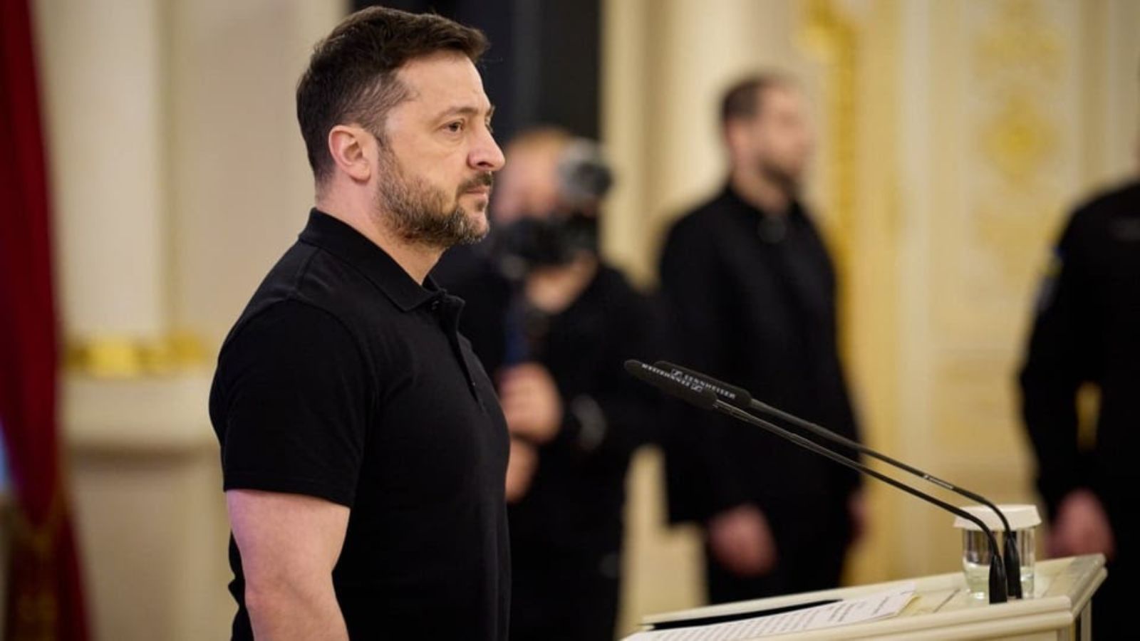 Profile photo of Volodymyr Zelenskyy standing near microphones in a hall. He is wearing a black polo shirt. Blurred figures of bodyguards are visible in the background.