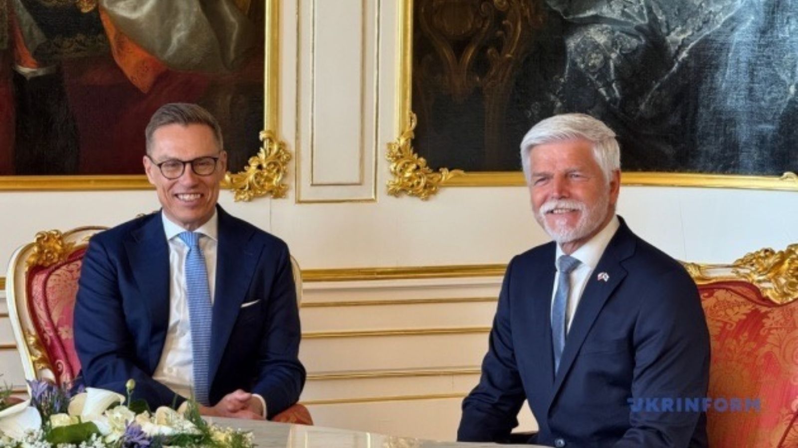 Alexander Stubb and Petr Pavel sitting at an ornate white table with gold detailing in a classical palace interior during an official meeting.