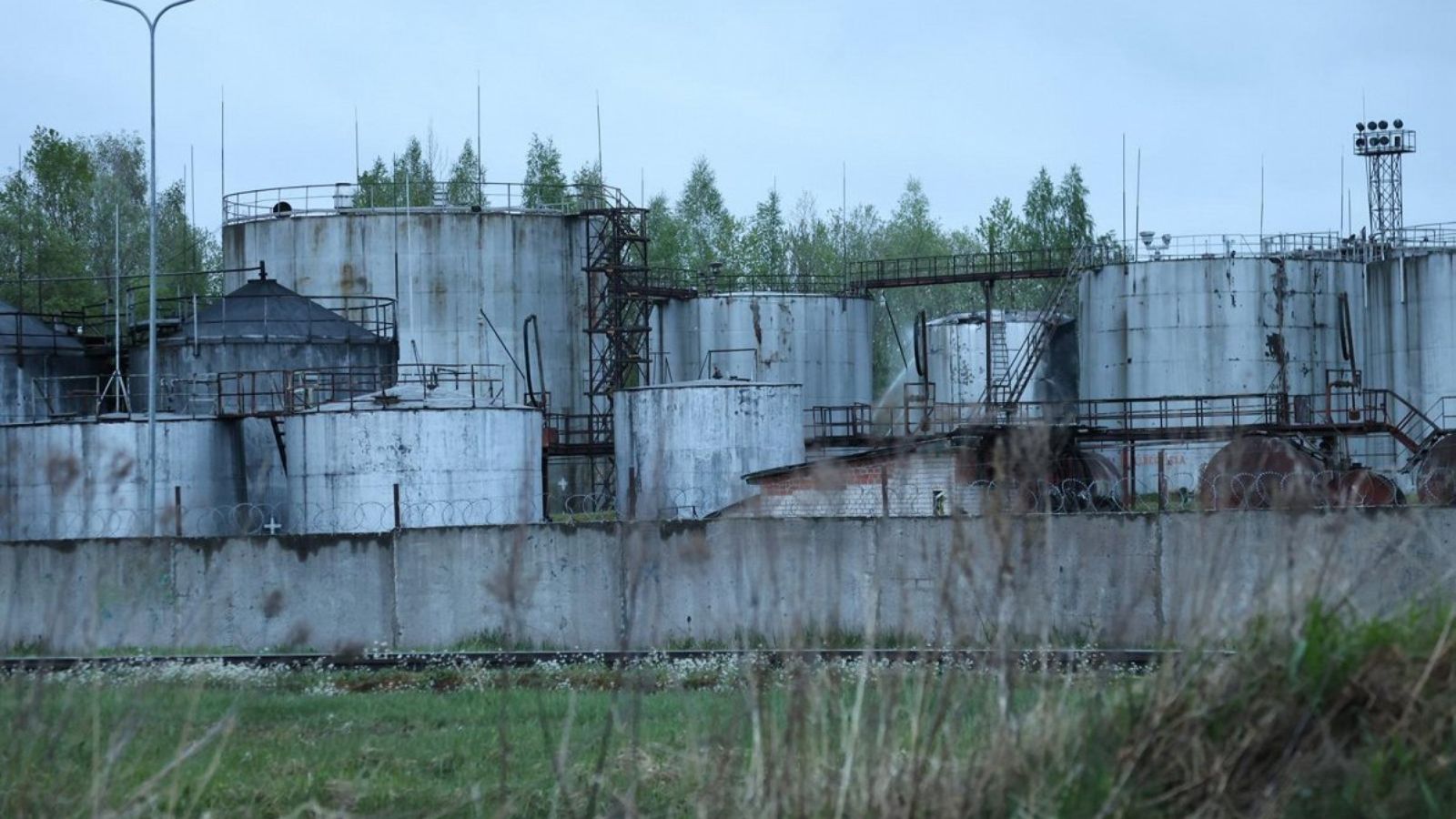 A wide shot of an industrial oil depot featuring several large cylindrical tanks behind a concrete fence topped with barbed wire.