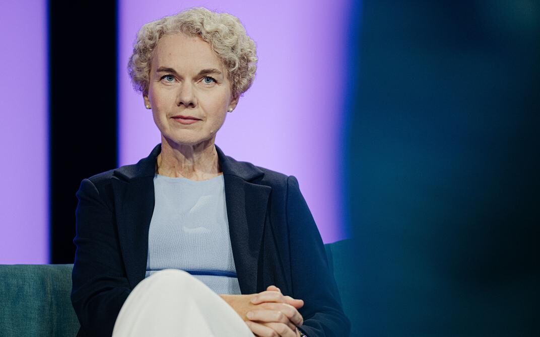 A medium shot of a woman with short, curly blonde hair sitting on a couch. She is wearing a dark navy blazer over a light blue top and white trousers, looking directly at the camera with a neutral expression against a soft purple background.
