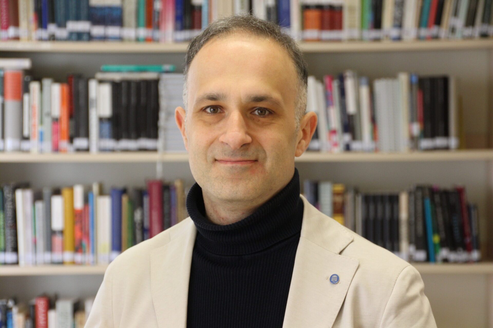 A medium shot of a man with short, thinning salt-and-pepper hair, wearing a black turtleneck and a light beige blazer. He is smiling slightly, posed against a blurred background of bookshelves filled with books.