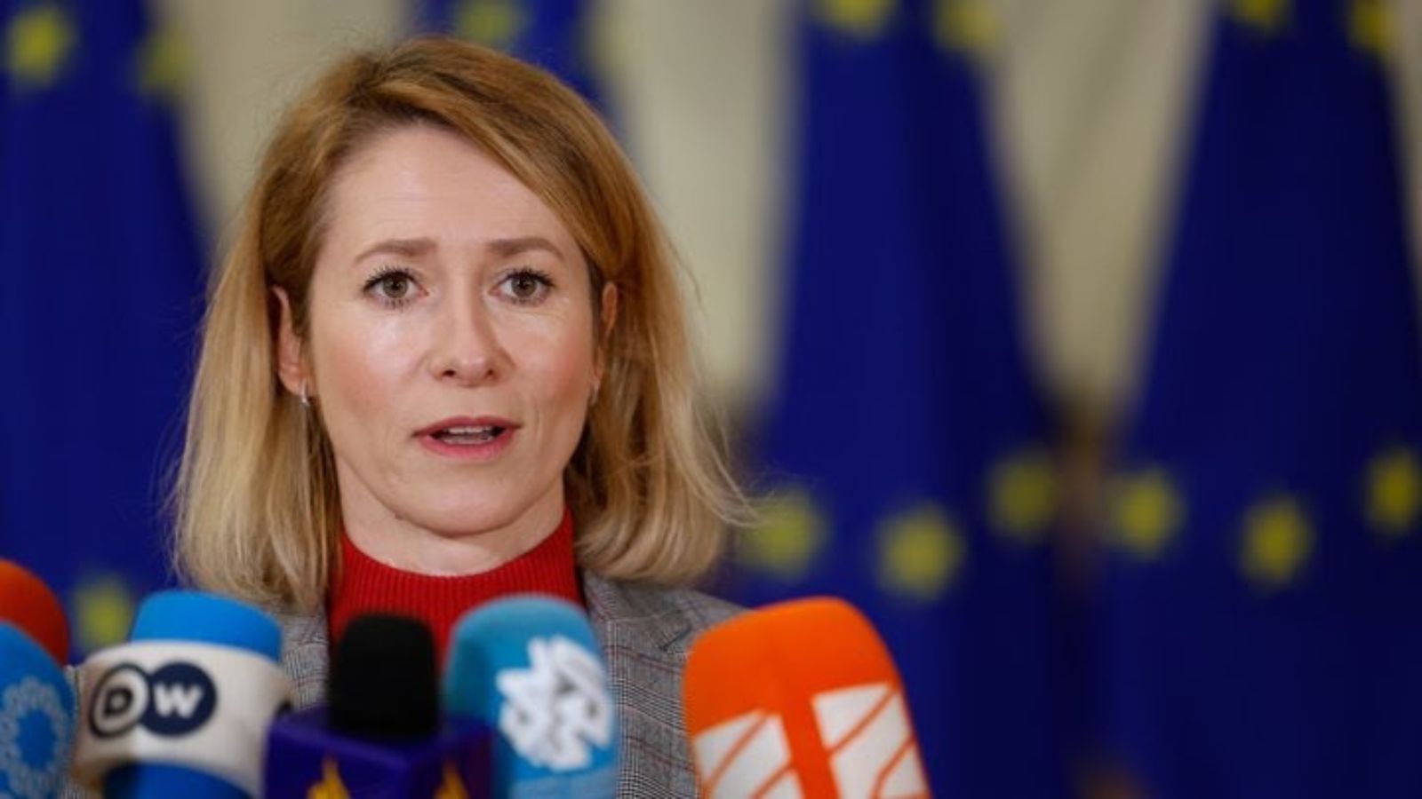 A close-up portrait of Kaja Kallas speaking into microphones. The European Union flags are blurred in the background.