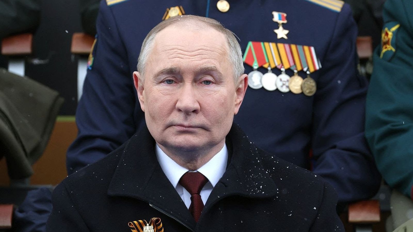 Close-up portrait of Vladimir Putin in a black coat with snow on his shoulders, with military officials in the background.