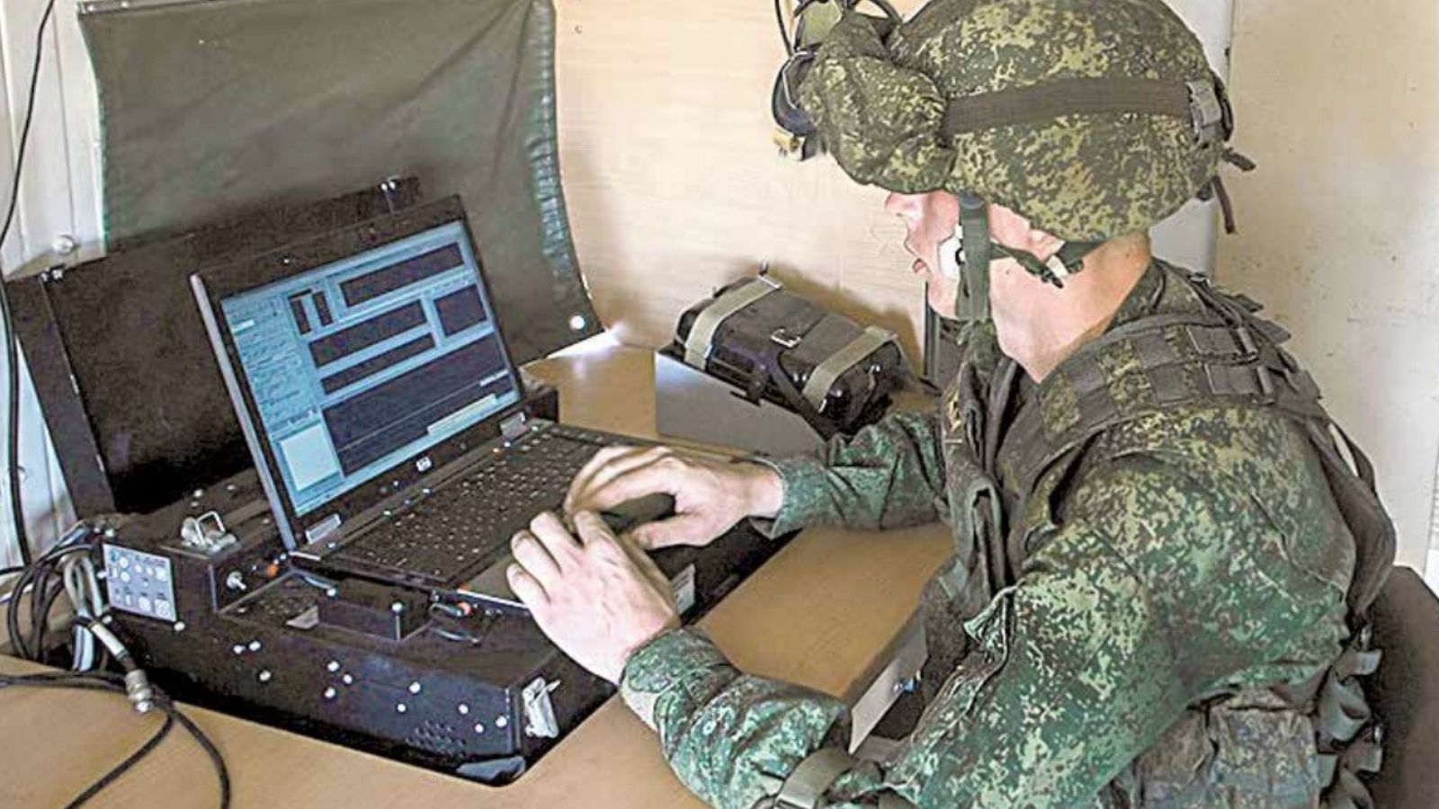 A Russian soldier in camouflage and a helmet operating a laptop in a field setting.