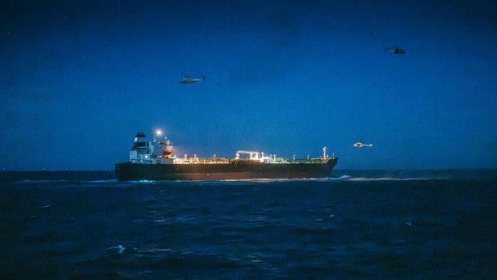 An oil tanker ship at night, with helicopters flying nearby, in the ocean.