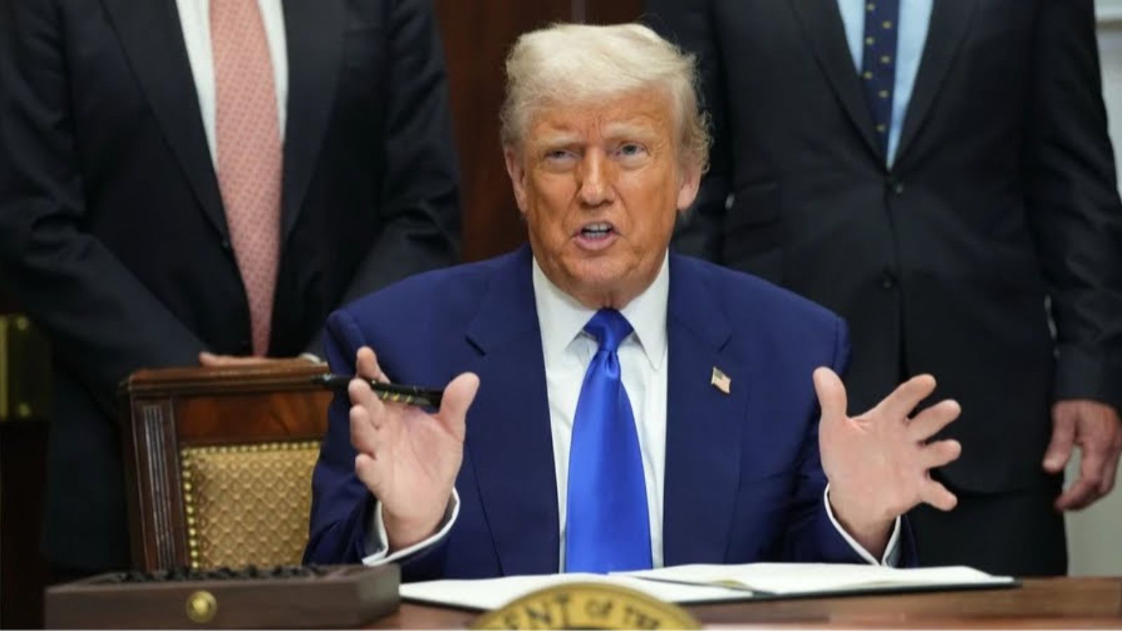 Donald Trump in a blue suit sitting at a desk and gesturing with his hands while speaking.