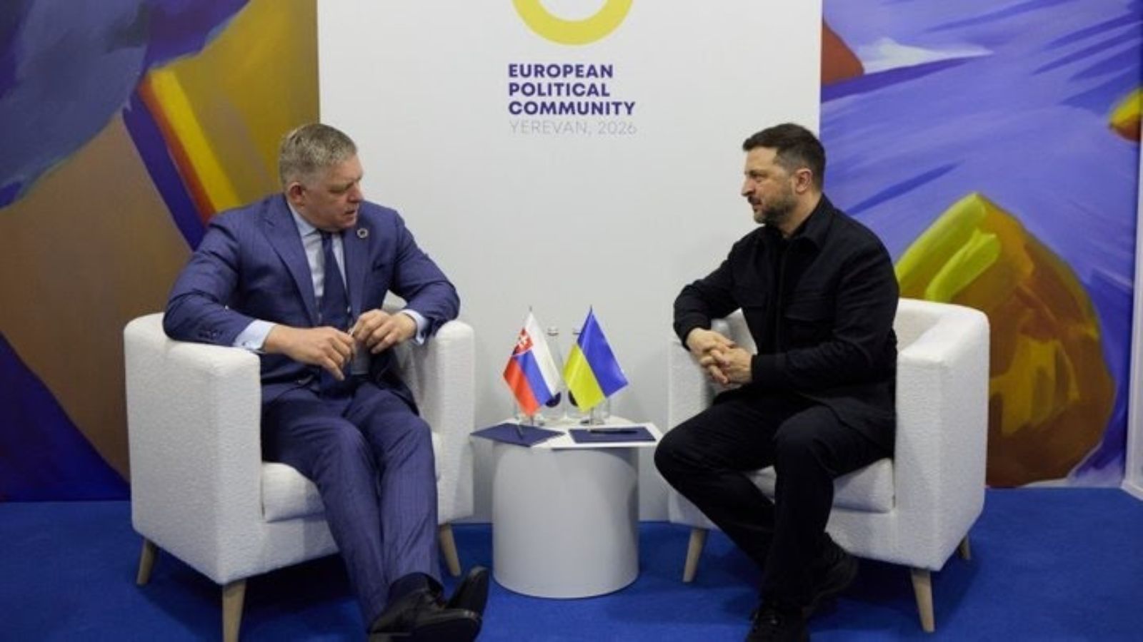 Ukrainian President Volodymyr Zelenskyy and Slovak Prime Minister Robert Fico sit opposite each other in white armchairs during a meeting. Small Slovak and Ukrainian flags sit on the table between them. The background is a brightly colored wall with a large sign that reads "EUROPEAN POLITICAL COMMUNITY YEREVAN, 2026".