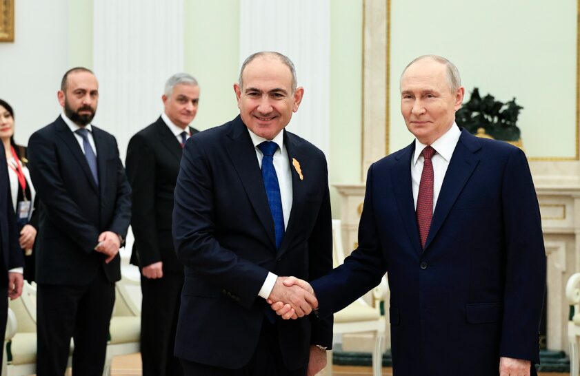 Armenian Prime Minister Nikol Pashinyan and Russian President Vladimir Putin shaking hands during an official meeting in a formal hall. Two other officials in suits stand in the background.